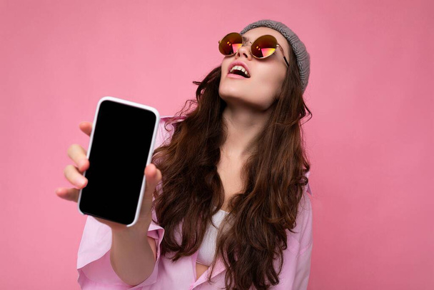 Mujer morena joven atractiva positiva con elegante camisa rosa sombrero gris y gafas de sol de colores aislados sobre fondo rosa sosteniendo en la mano y mostrando el teléfono móvil con pantalla vacía para - Foto, imagen