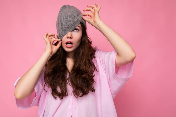 Foto záběr okouzlující bláznivé emocionální mladá bruneta žena stojící izolované přes růžové pozadí zdi na sobě růžová košile a šedý klobouk a baví - Fotografie, Obrázek