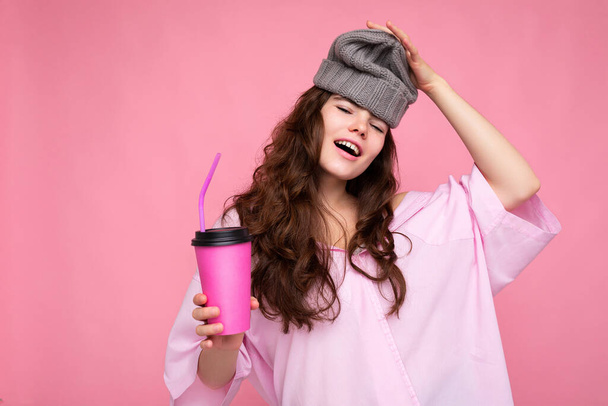 Hermosa joven morena positiva rizado mujer con camisa de color rosa y sombrero gris aislado sobre fondo rosa sosteniendo taza de café de papel para mockup beber y divertirse - Foto, imagen