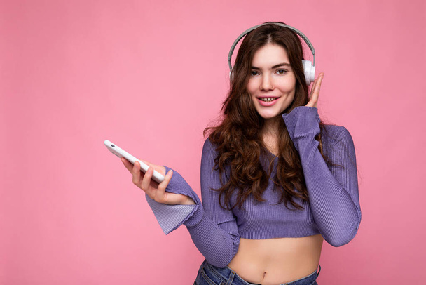 Photo of beautiful joyful smiling young woman wearing stylish casual clothes isolated over background wall holding and using mobile phone wearing white bluetooth headphones listening to music and - Fotografie, Obrázek