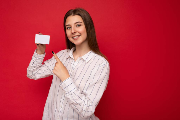 Fénykép lövés vonzó pozitív mosolygós fiatal sötét szőke nő visel fehér blúz elszigetelt piros háttér gazdaság hitelkártya nézi kamera ujjal mutogató műanyag érintésmentes kártya - Fotó, kép
