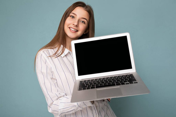 Közelkép portré gyönyörű mosolygós fiatal nő gazdaság netbook számítógép nézi kamera viselt fehér ing elszigetelt kék háttér - Fotó, kép