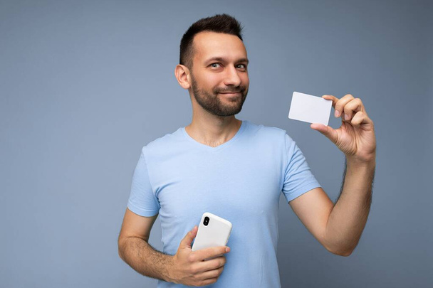 Φωτογραφία του χαρούμενου όμορφου νεαρού αξύριστου brunet άνδρα με γενειάδα που φοράει καθημερινά μπλε t-shirt απομονωμένο σε μπλε φόντο κρατώντας και χρησιμοποιώντας κινητό τηλέφωνο και πιστωτική κάρτα κάνοντας την πληρωμή κοιτάζοντας κάμερα - Φωτογραφία, εικόνα