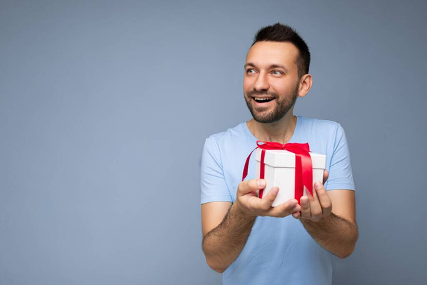 Photo de belle brune positive jeune homme non rasé isolé sur fond bleu mur portant t-shirt bleu tenant boîte cadeau blanche avec ruban rouge et regardant sur le côté - Photo, image