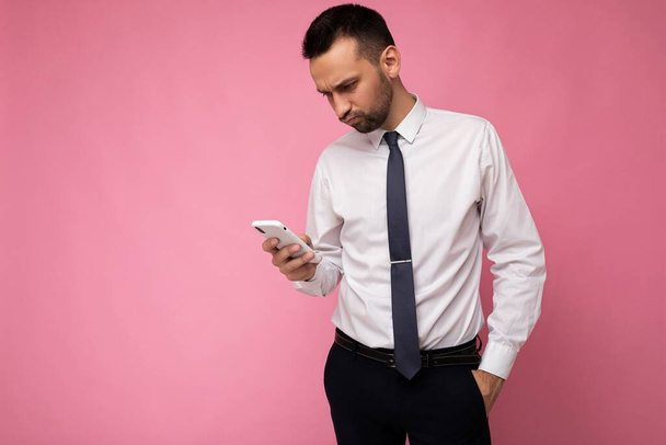 Photo shot of handsome concentrated good looking businessman wearing casual stylish outfit poising isolated on background with empty space holding in hand and using mobile phone messaging sms looking - Photo, Image