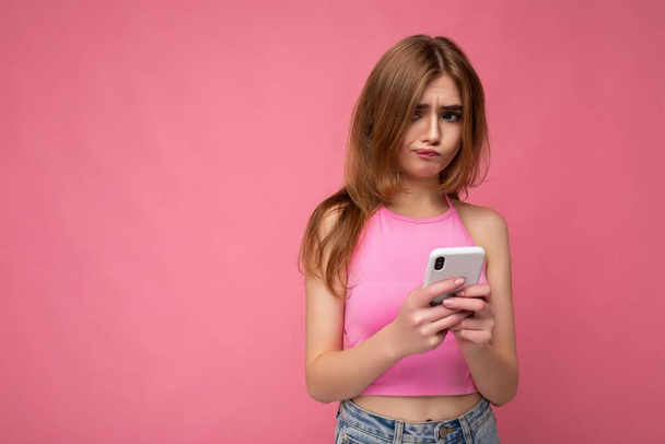 Closeup photo of beautiful upset thoughtful young blonde woman wearing pink top poising isolated on pink background with empty space holding in hand and using mobile phone surfing on the internet - Photo, Image