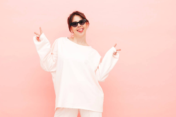Young beautiful smiling female in trendy summer hipster clothes. Sexy carefree woman posing near pink wall in studio. Positive model having fun indoors. Cheerful and happy.Shows peace sign and tongue - Foto, afbeelding