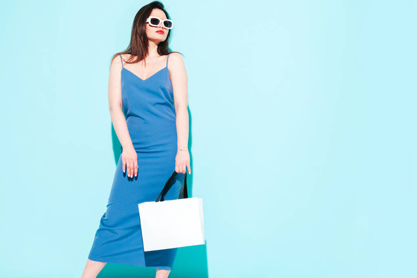 High fashion portrait of young beautiful brunette woman wearing nice blue summer dress.Sexy trendy model posing near blue wall in studio.Fashionable female in sunglasses. Holding shopping bag - Foto, immagini