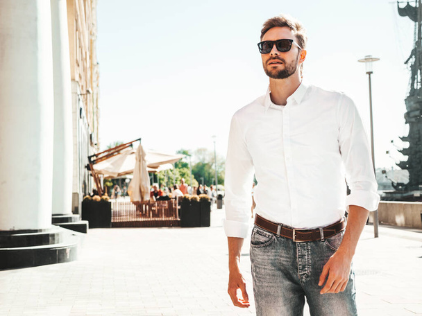 Portrait of handsome smiling stylish hipster lambersexual model.Modern man dressed in white shirt. Fashion male posing on the street background in sunglasses. Outdoors at sunset  - Фото, изображение