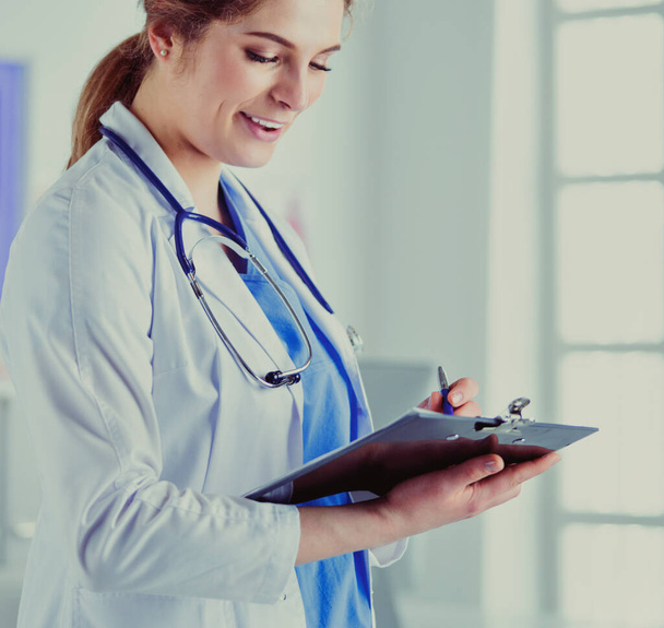Doctor with a stethoscope, holding a notebook in his hand. Close-up of a female doctor filling up medical form at clipboard while standing straight in hospital - Foto, imagen