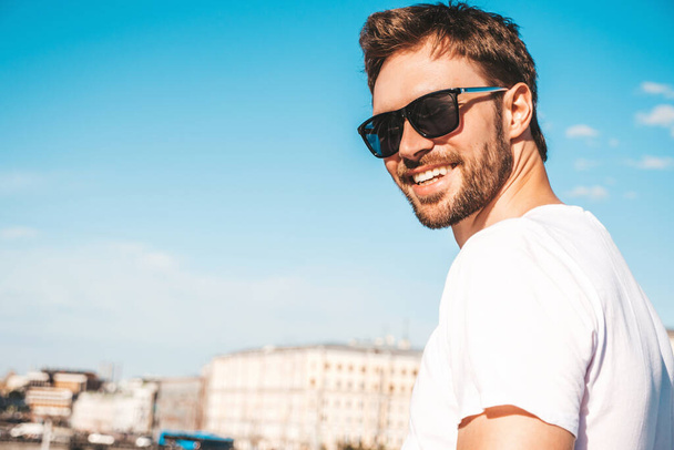 Closeup portrait of handsome smiling  hipster lambersexual model.Stylish man dressed in white T-shirt. Fashion male posing behind blue sky on the street background in sunglasses - Valokuva, kuva