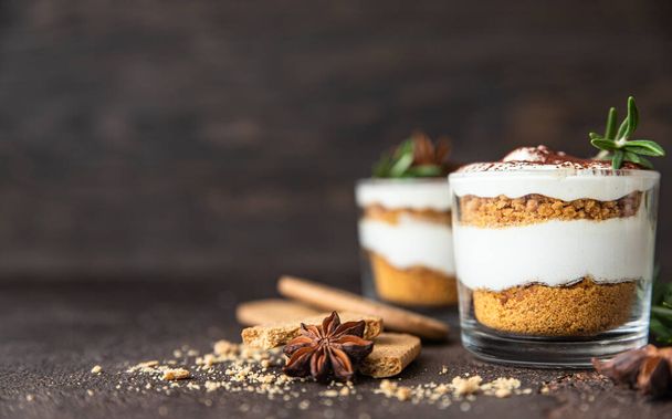 Layered dessert in glass jar with cookie crumble and whipped cream decorated with rosemary and anise, dark brown background. No bake cheesecake, trifle or pudding. - 写真・画像