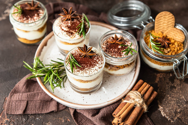 Layered dessert in glass jar with cookie crumble and whipped cream decorated with rosemary and anise, dark brown background. No bake cheesecake, trifle or pudding. - Foto, Imagem