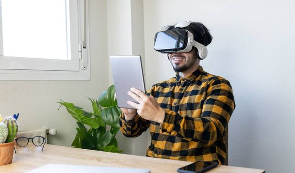 sentado hombre guapo utiliza gafas de realidad virtual mientras sostiene una tableta delante de él - Foto, imagen