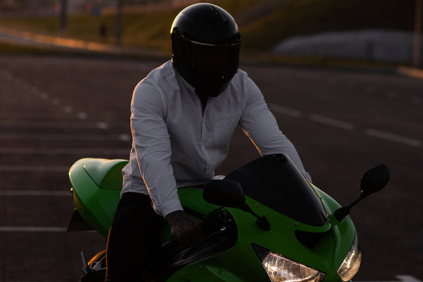 Неузнаваемый стильный мотоциклист в шлеме сидит на припаркованном мотоцикле в городе на фоне закатного неба - Фото, изображение