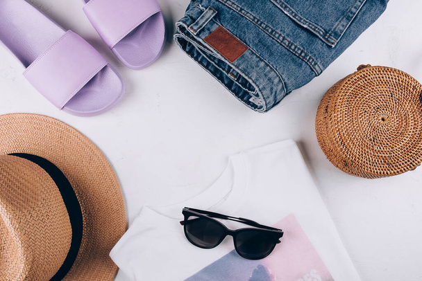 Overhead summer still life. Casual women's clothing, acccessories. T-shirt, slippers, shorts, organic rattan bag, straw hat, sunglasses. Vacation, travel concept. Top view flat lay - Photo, Image