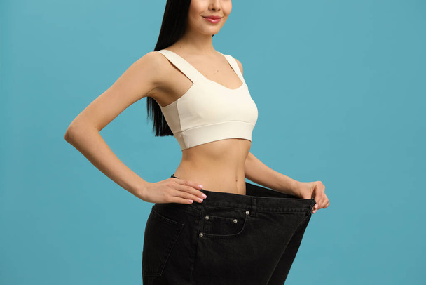 Young woman wearing big jeans after weight loss on turquoise background, closeup - Foto, Bild