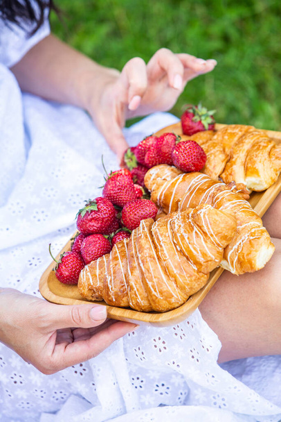Outdoors picnic in a lush green park with a tasty croissant, fruits, donuts and wine on grass. Summer picnic on the blanket. - Photo, Image