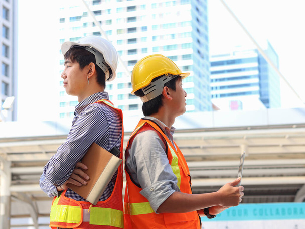 Портрет двох розумних молодих азіатських інженерів, одягнених у безпечний жилет і шолом, співробітники конфіденційно стоять за межами офісу в центрі міста з нечітким тлом будівлі хмарочоса. - Фото, зображення