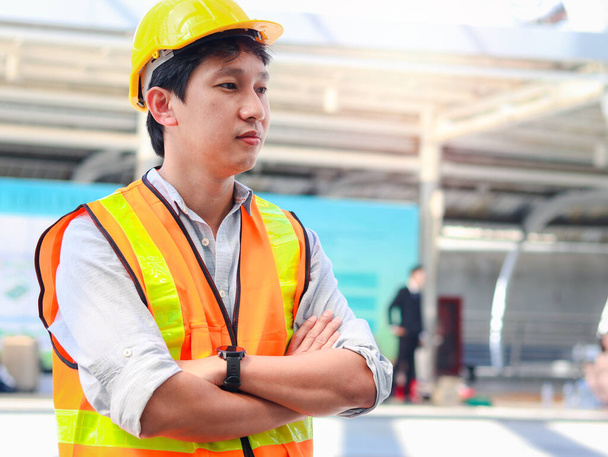 Venkovní portrét asijského mužského inženýra v bezpečnostní vestě a helmě stojící se zkříženýma rukama v moderním městě, mladý muž pracující na ulici venku.  - Fotografie, Obrázek