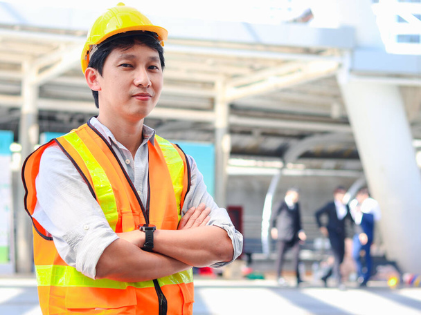 Zewnętrzny portret azjatyckiego inżyniera w kamizelce bezpieczeństwa i kasku stojącego z rękami skrzyżowanymi w nowoczesnym mieście, młodego człowieka idącego ulicą na zewnątrz.  - Zdjęcie, obraz
