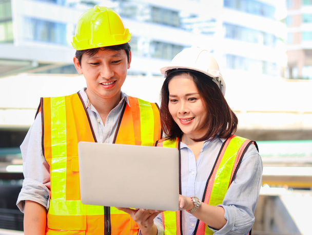 Két fiatal ázsiai férfi és női mérnök biztonsági mellényt és sisakot visel, laptopot használva, együtt beszélgetnek a projektről, miközben a belvárosi irodán kívül sétálnak..  - Fotó, kép