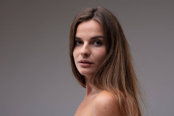 Natural portrait of a sincere young woman with naked shoulders looking back over her shoulder at the camera with wide eyes and parted lips in a head and shoulders portrait against a grey studio background - Foto, imagen