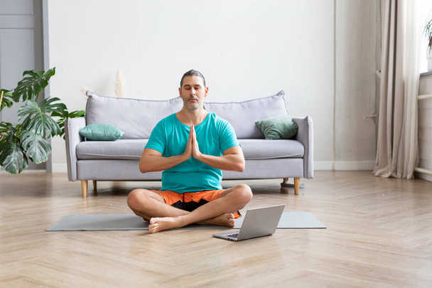 Фотографія чоловіка медитує перед монітором ноутбука. Він сидить в положенні лотоса в домашньому декорі. Молитовна поза
. - Фото, зображення