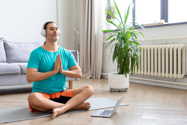 Um homem medita na frente de um monitor de laptop. Ele se senta na posição de lótus na decoração da casa. Postura de oração. - Foto, Imagem