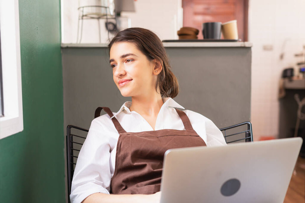 Beautiful caucasian barista woman sits blankly near window and looks outside coffee shop while using laptop in breaking time. Barista work at coffee bar and food service business - Φωτογραφία, εικόνα