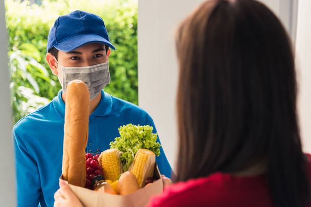 Asian young delivery man in uniform wear protective face mask he making grocery service giving fresh food to woman customer receiving front house under pandemic coronavirus, Back to new normal concept - Foto, Imagem
