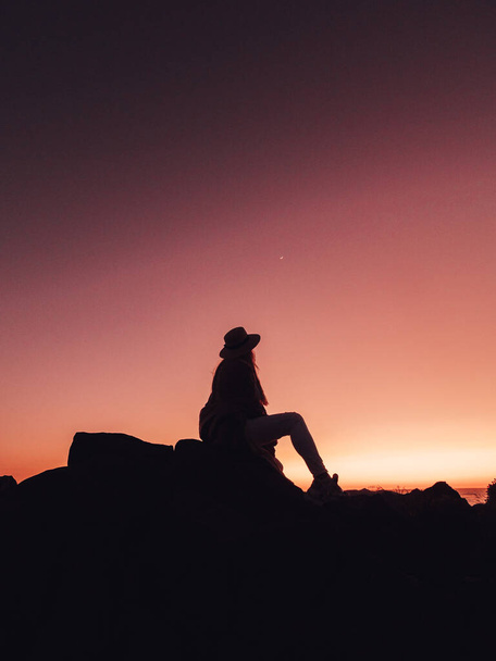a silhouette of a female sitting on the stone and looking at sunset under a pink sky - Photo, image