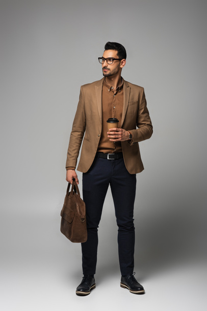 Hombre de negocios musulmán en chaqueta marrón sosteniendo taza de papel y bolso sobre fondo gris - Foto, imagen