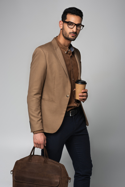Stilvoller muslimischer Geschäftsmann hält Pappbecher und Handtasche isoliert auf grau  - Foto, Bild