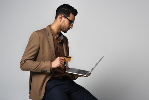 Side view of arabian businessman holding credit card and laptop isolated on grey  - Φωτογραφία, εικόνα