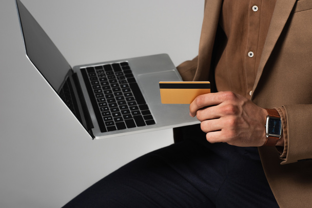 Vue recadrée d'un homme d'affaires en veste brune tenant une carte de crédit et un ordinateur portable flou isolé sur gris  - Photo, image