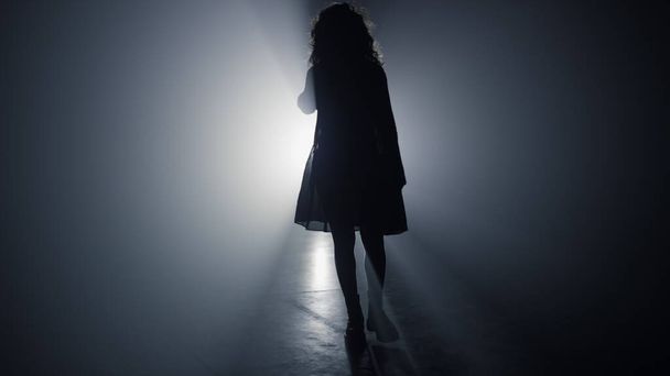 Женщина в силуэте ходит в темноте. Девушка идет в прожекторе фоне. - Фото, изображение
