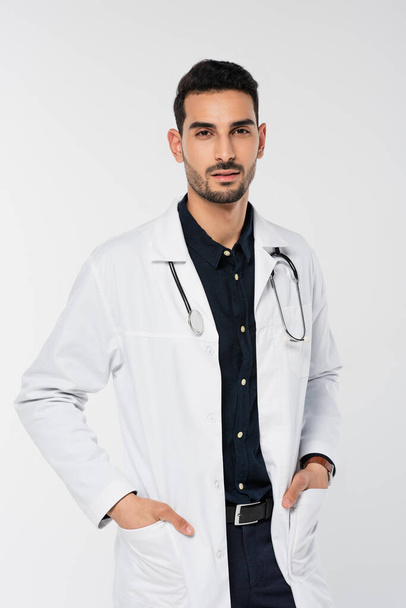 Арабский врач со стетоскопом, держащийся за руки в карманах белого халата, изолированного на сером  - Фото, изображение