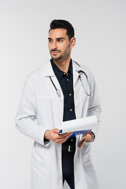 Muslim doctor in white coat holding clipboard and looking away isolated on grey  - Fotoğraf, Görsel