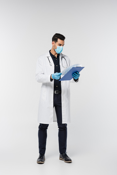 Médico árabe en máscara médica sujetando portapapeles sobre fondo gris  - Foto, Imagen