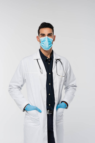 Arabischer Arzt mit medizinischer Maske und Latex-Handschuhen blickt isoliert auf graue Kamera  - Foto, Bild