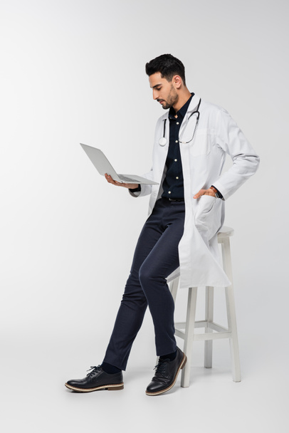 Vista laterale del medico arabo utilizzando laptop vicino sedia su sfondo grigio  - Foto, immagini