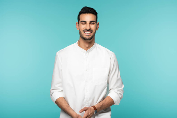 Άραβας άνδρας με λευκό πουκάμισο χαμογελά στην κάμερα απομονωμένος στο μπλε  - Φωτογραφία, εικόνα