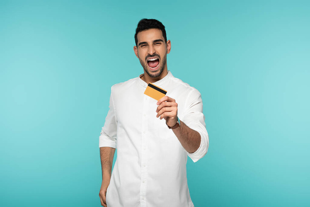 Hombre árabe excitado mostrando tarjeta de crédito aislada en azul  - Foto, Imagen