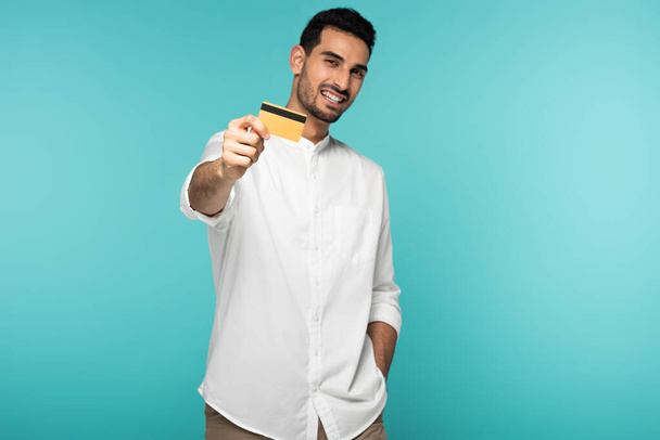 Bulanık Arap adamın elindeki kredi kartı mavi üzerine izole bir şekilde gülümsüyor.  - Fotoğraf, Görsel