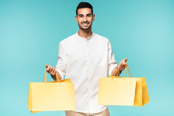 Positive arabian man holding yellow shopping bags isolated on blue  - Photo, Image