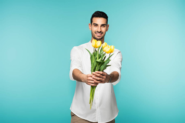 Smiling arabian man holding yellow flowers isolated on blue  - Foto, Imagen