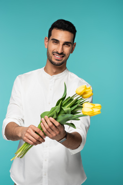 Yellow tulips in hands of smiling arabian man on blurred background isolated on blue  - Zdjęcie, obraz