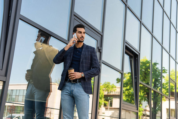 Hombre de negocios árabe hablando en smartphone y sosteniendo café para salir al aire libre  - Foto, Imagen