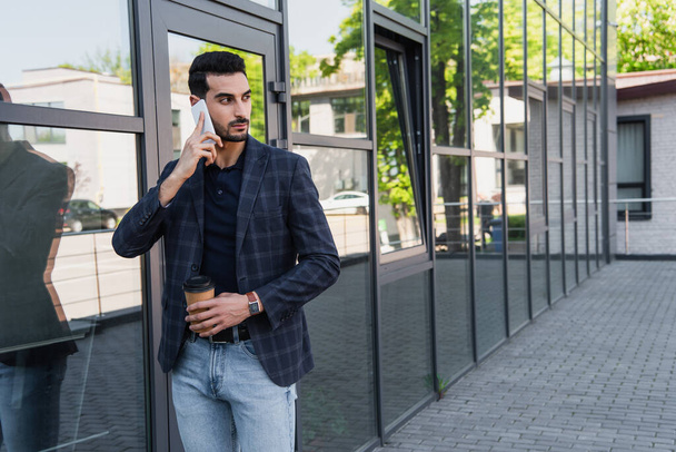 Beau gestionnaire musulman avec tasse en papier parlant sur smartphone à l'extérieur  - Photo, image